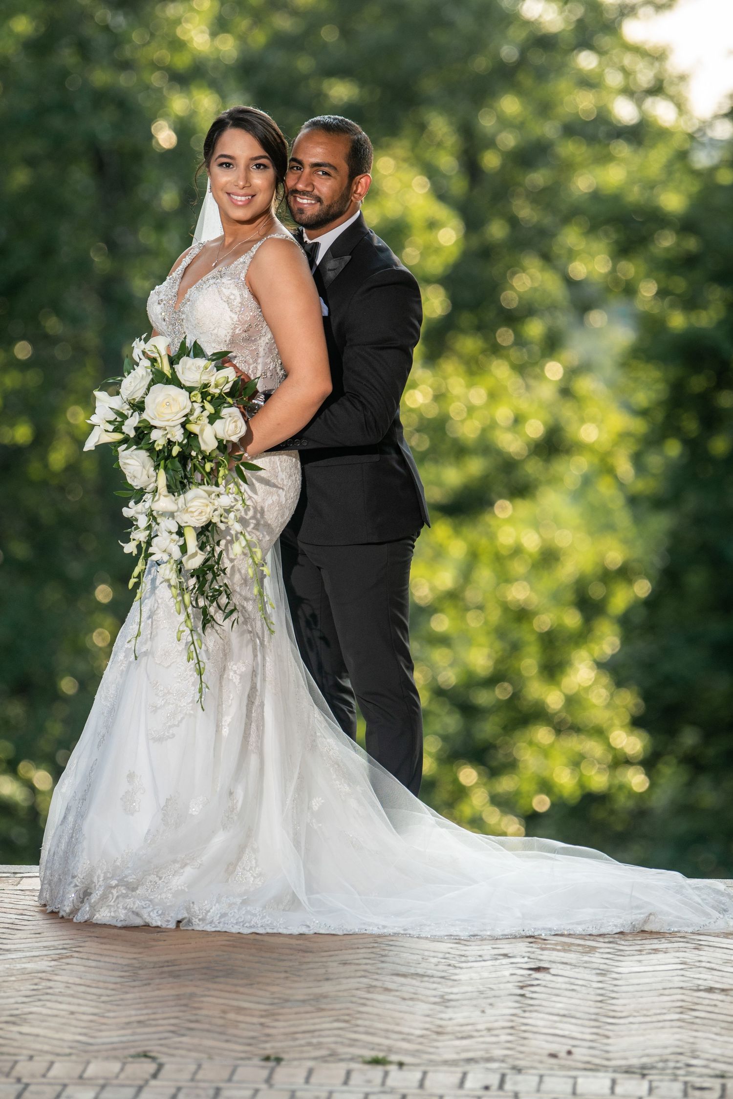 Wedding couple posing with white floral bouquet against lush green summer background.