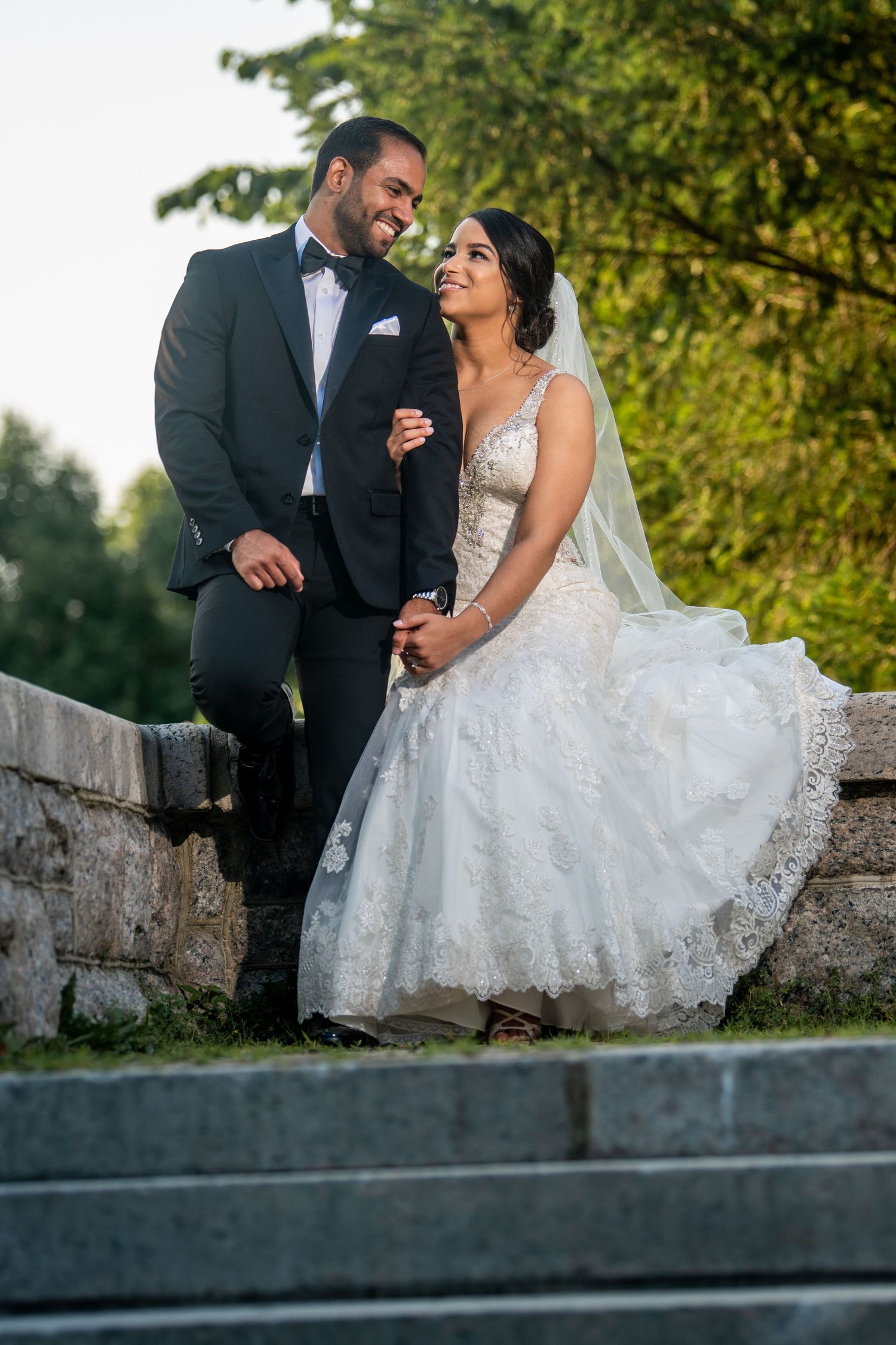 Newlyweds sharing an intimate moment on a stone wall surrounded by green foliage.