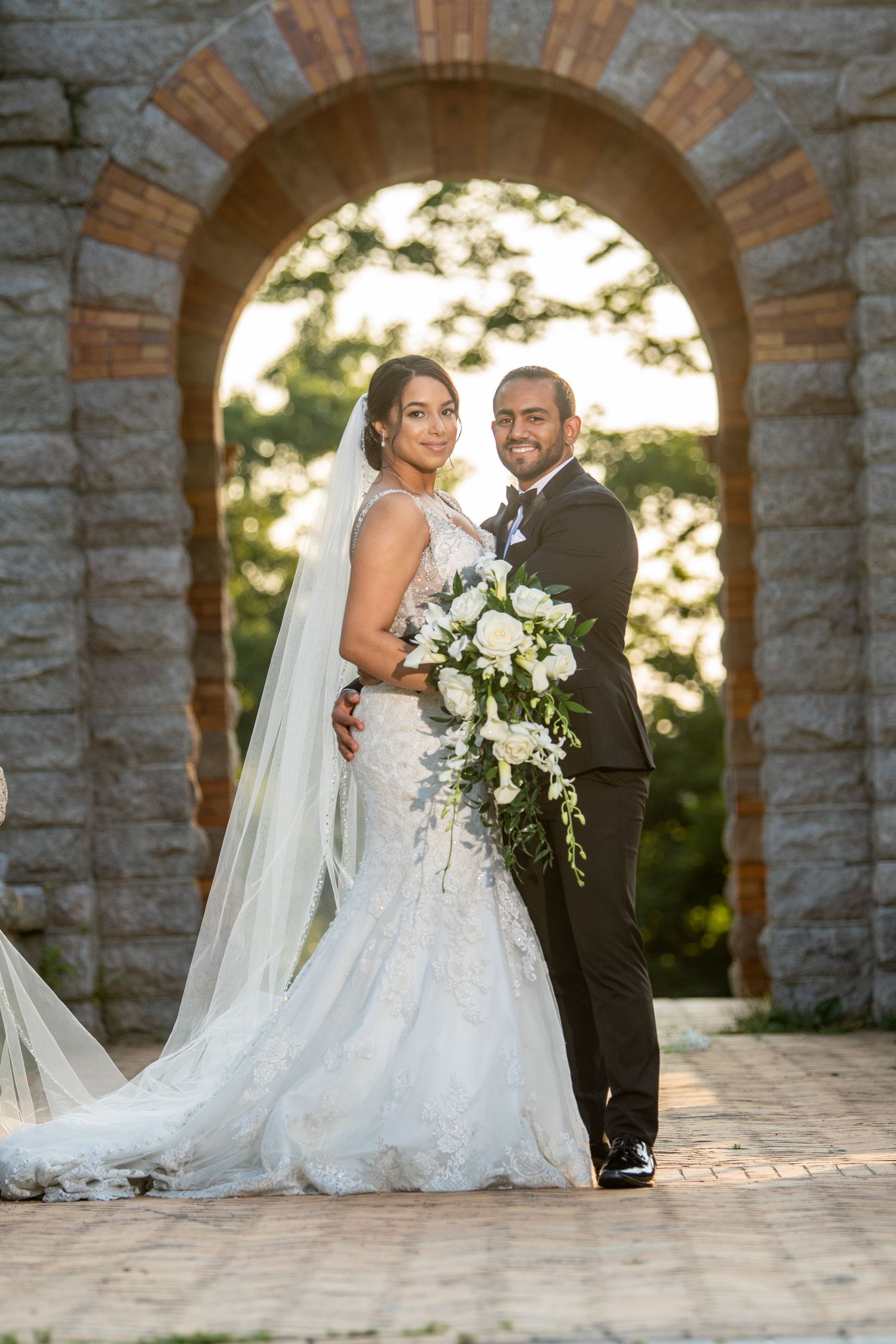 Wedding couple poses under stone archway during golden hour sunset.