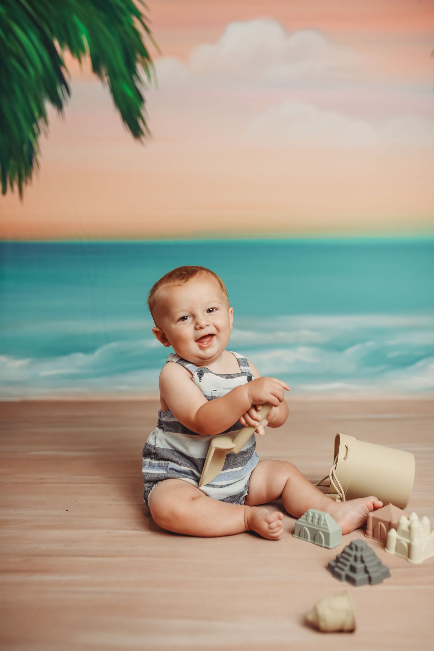 Baby in striped outfit plays with sand toys on beach backdrop with turquoise water and palm trees.