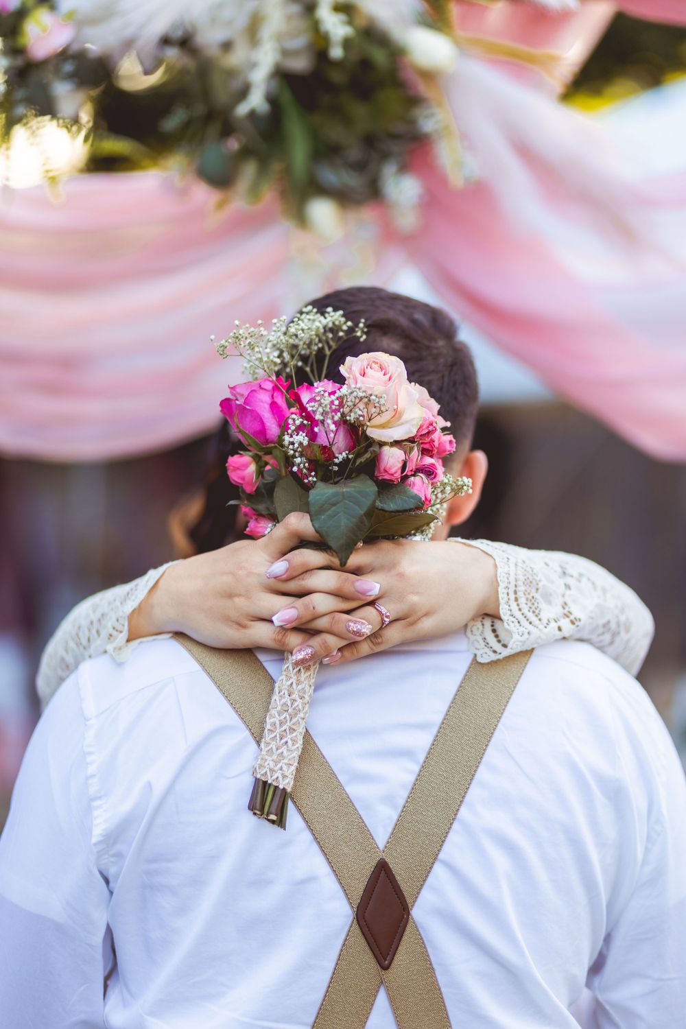 Bride embracing groom from behind holding pink rose bouquet, groom wearing suspenders at wedding.