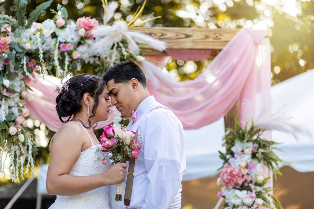 Bride and groom touching foreheads under floral arch with pink draping at outdoor wedding.