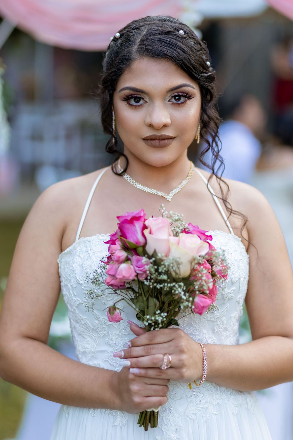 Beautiful bride in white lace gown holding pink rose bouquet at outdoor wedding ceremony.