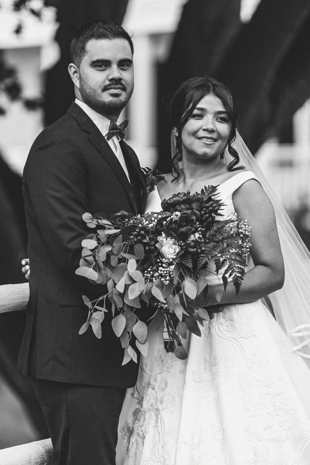 Black and white photo of a bride and groom posing together, bride holding a lush floral bouquet with greenery.