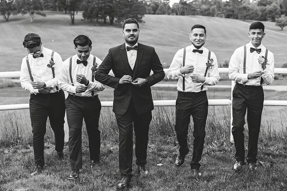 Black and white photo of a groom and groomsmen in suits and bow ties posing outdoors near a white fence.