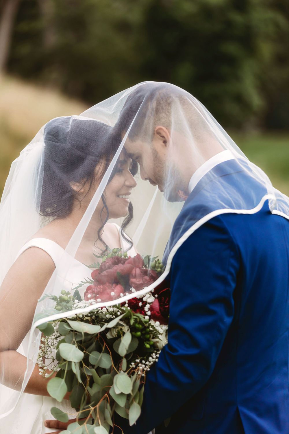 Bride and groom touching foreheads under a flowing veil, bride holding a red and green bouquet outdoors.
