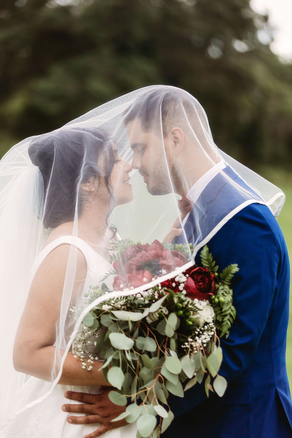 Bride and groom in blue suit touching foreheads under a veil, holding a red and white floral bouquet.