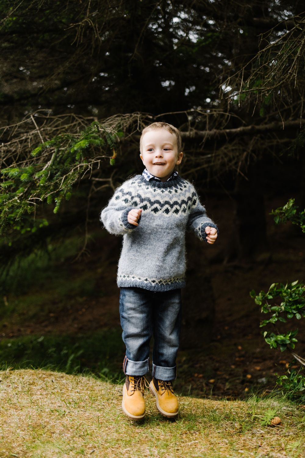 Young child jumping while wearing gray patterned sweater and jeans in forest setting.