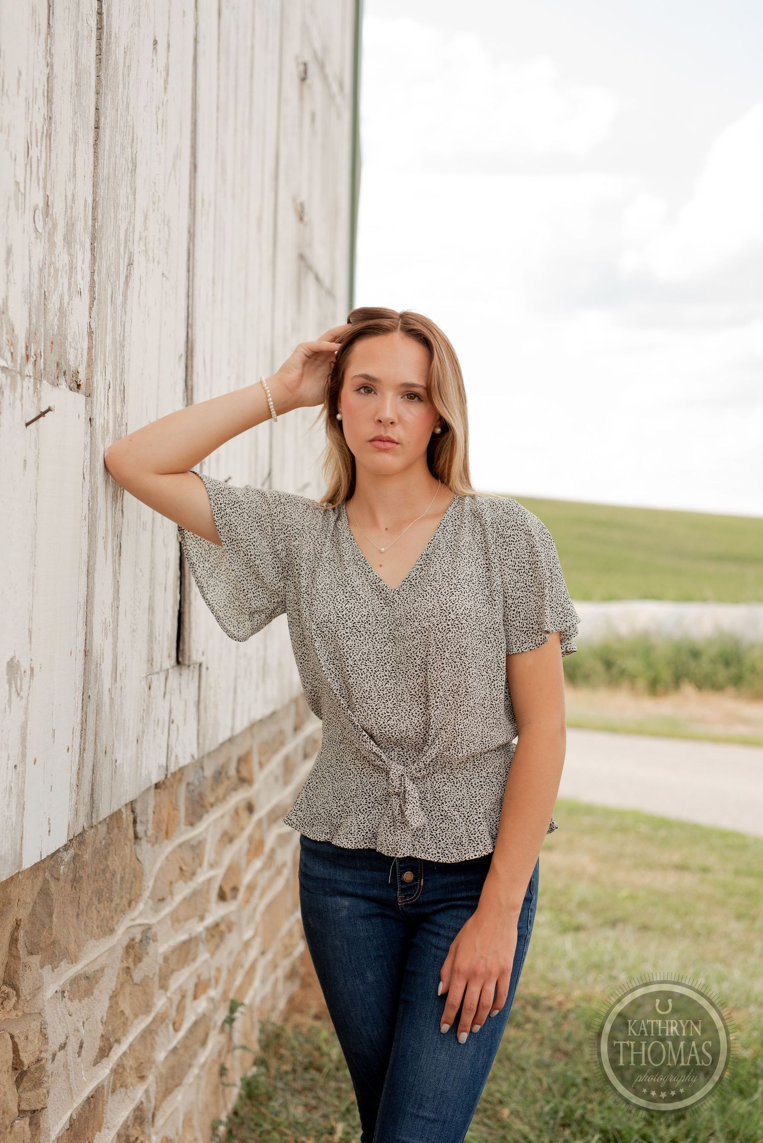 A person in a patterned blouse poses against a weathered white barn wall in natural lighting.