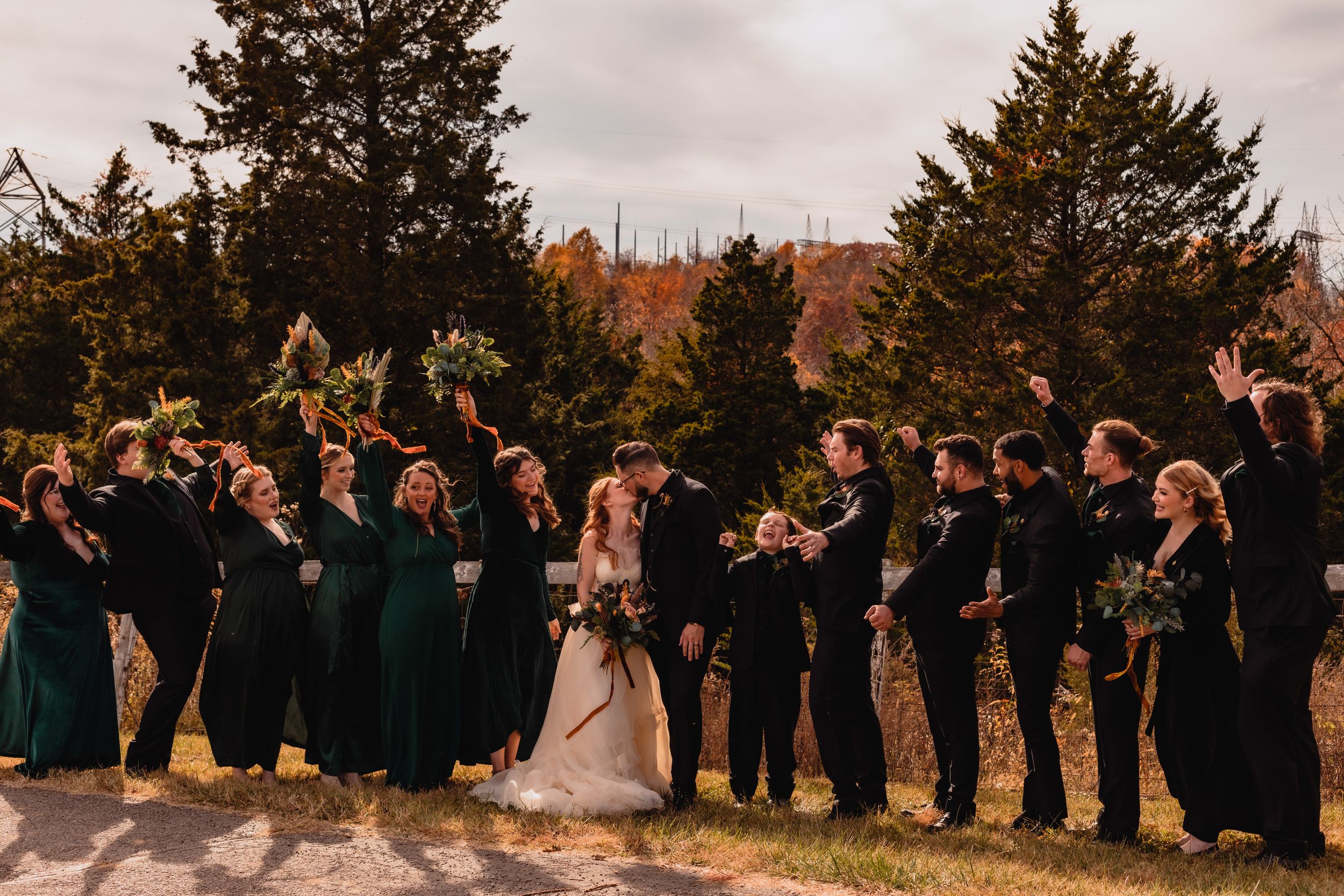 Large wedding party lined up outdoors with evergreen trees and cloudy sky in the background.