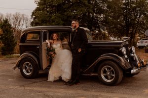 Vintage black car serves as backdrop for wedding photos in rustic outdoor setting.