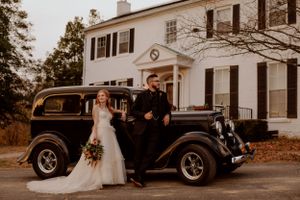 A wedding party poses with a classic black vintage car outside a white colonial-style house.