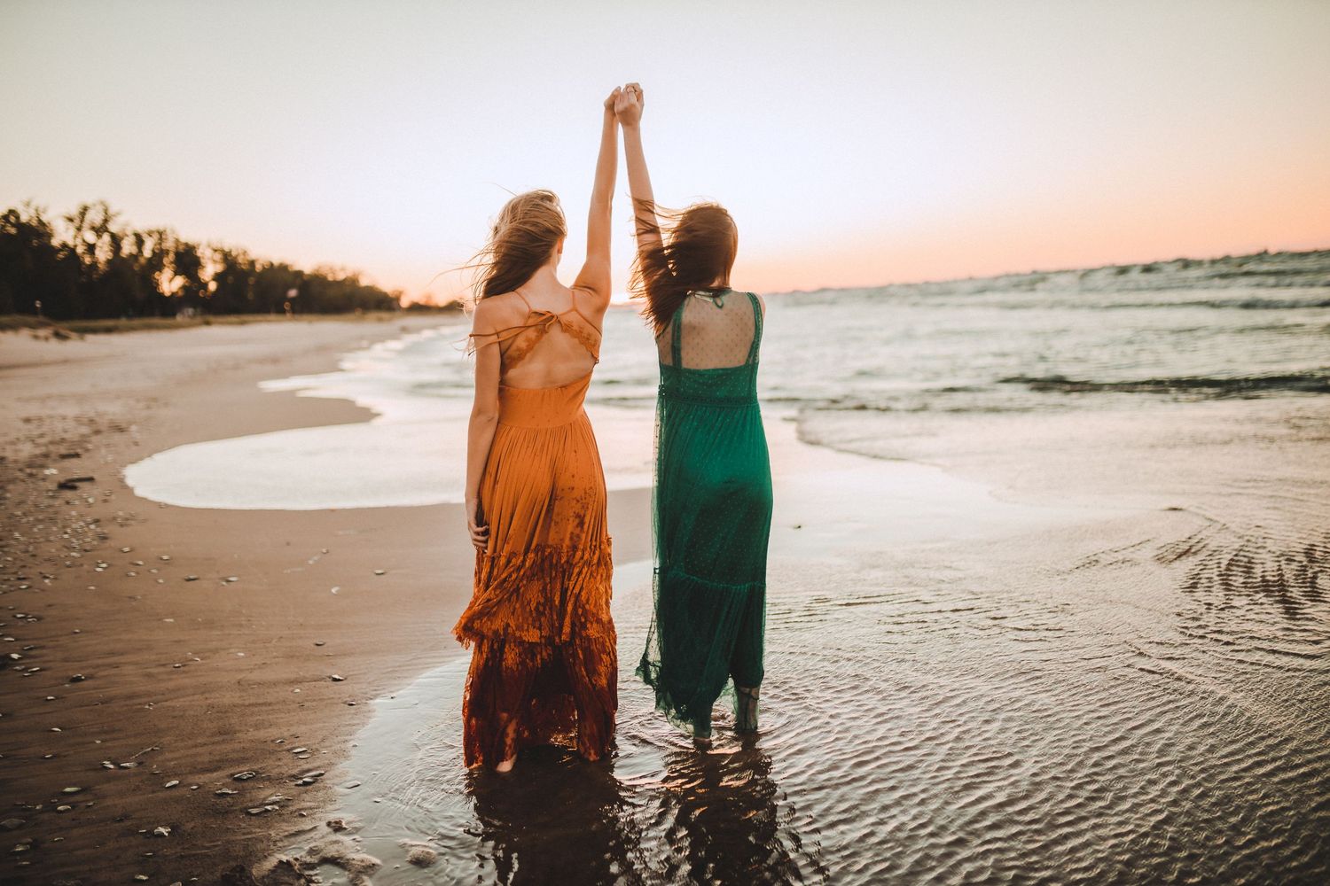 Two women in flowing dresses holding hands at sunset on a beach.
