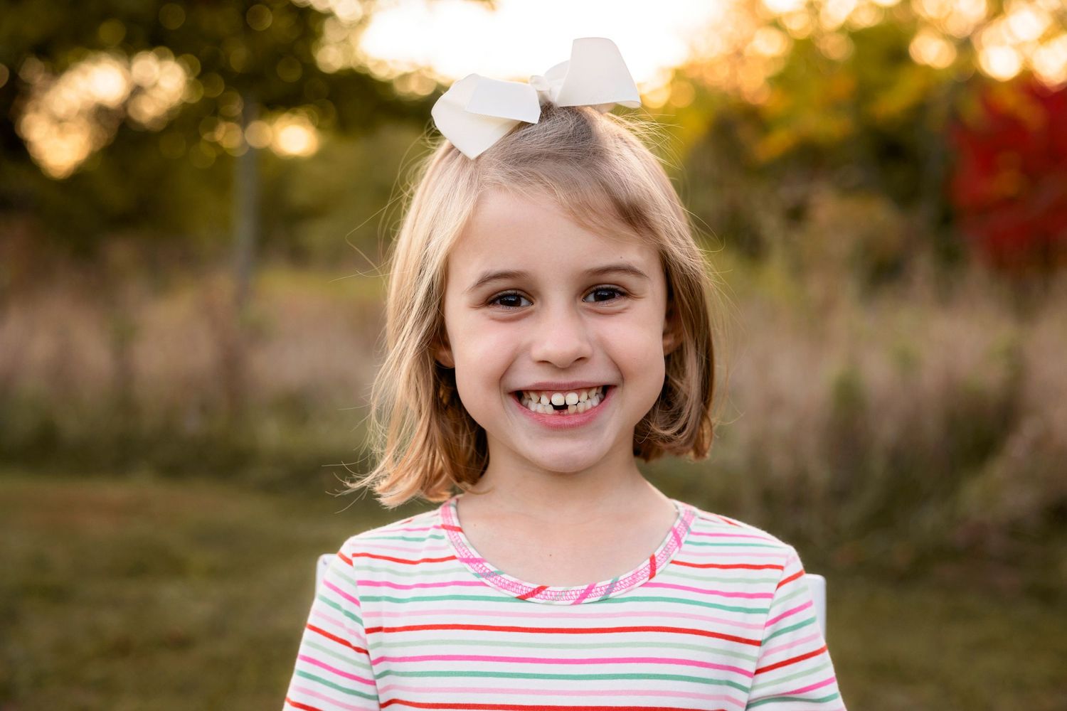 Child in striped shirt smiles brightly outdoors during golden hour.