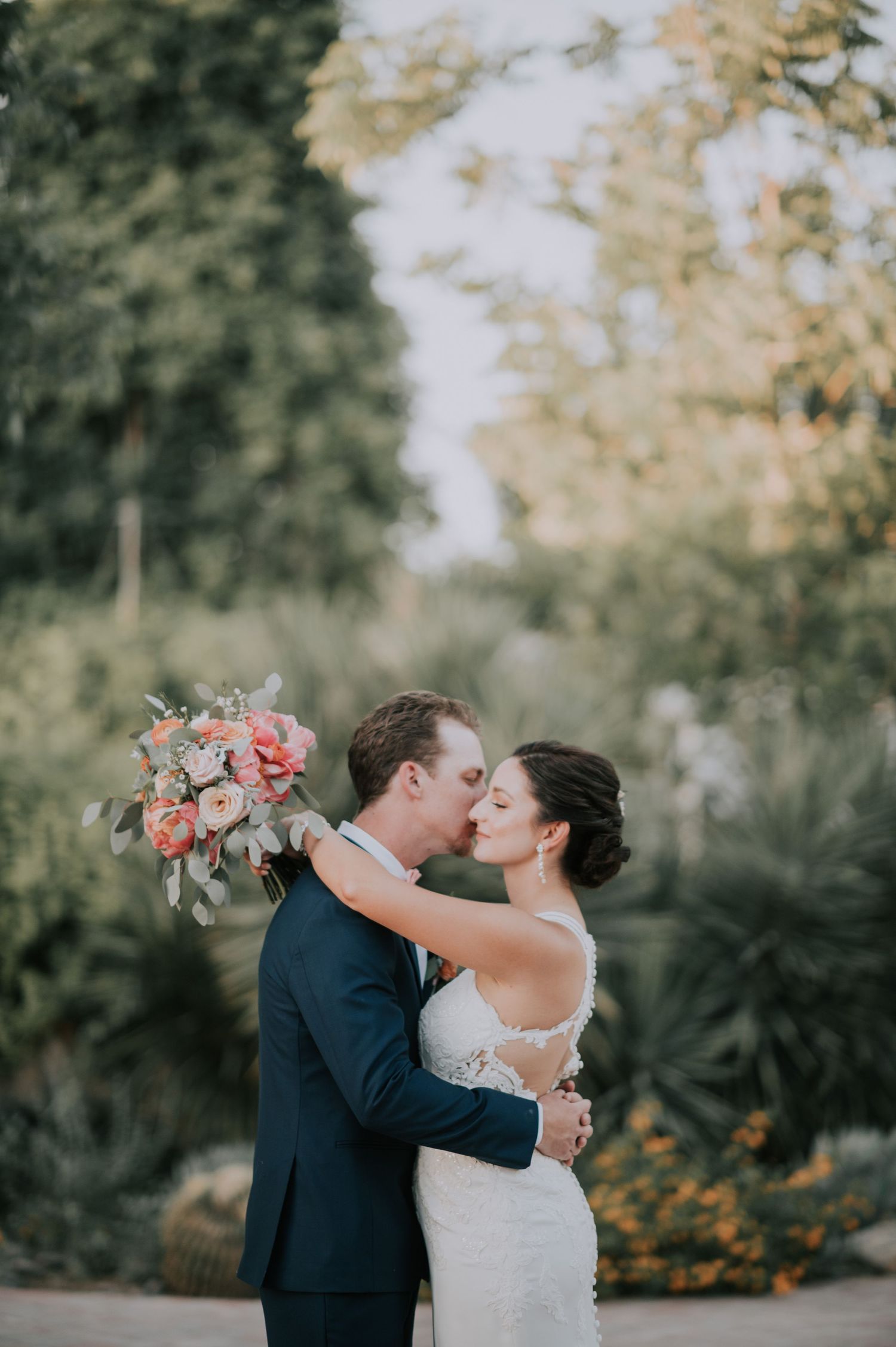 A couple shares a romantic moment in a garden with coral floral bouquet.