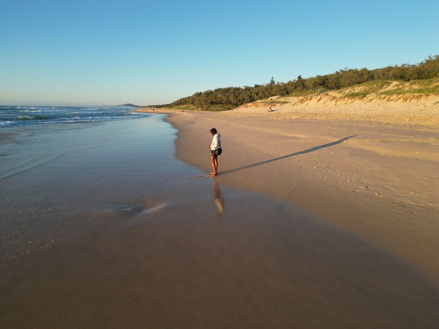 Lone figure walking along empty beach at sunset with waves and sand dunes.