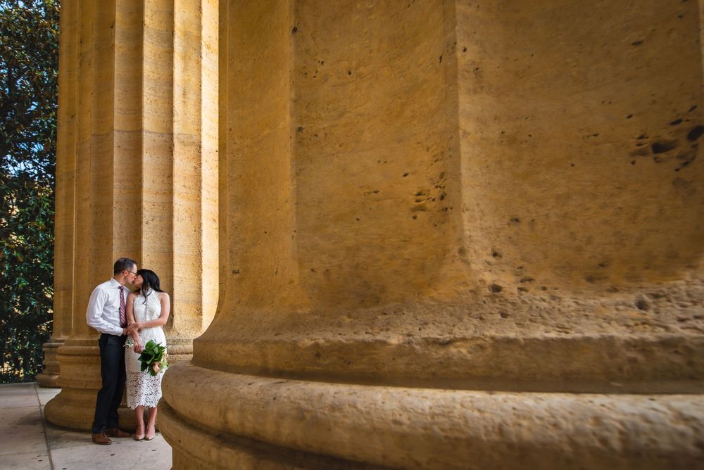 A wedding couple stands between elegant stone columns casting dramatic shadows at a historic venue.