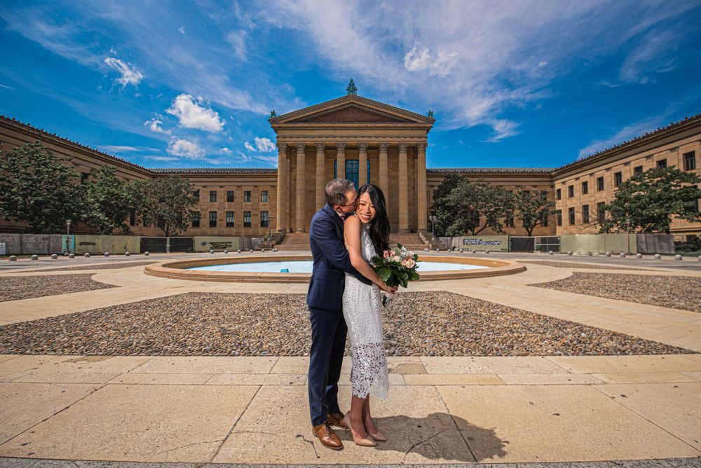 Couple poses for wedding photo in front of Philadelphia Museum of Art on sunny day.