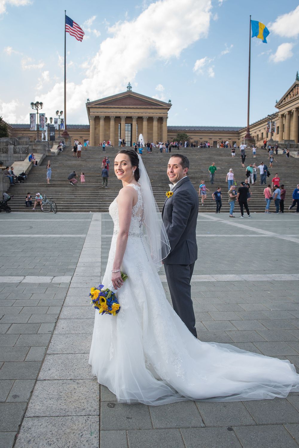 A wedding couple poses on the Philadelphia Museum of Art steps with American flags flying above.