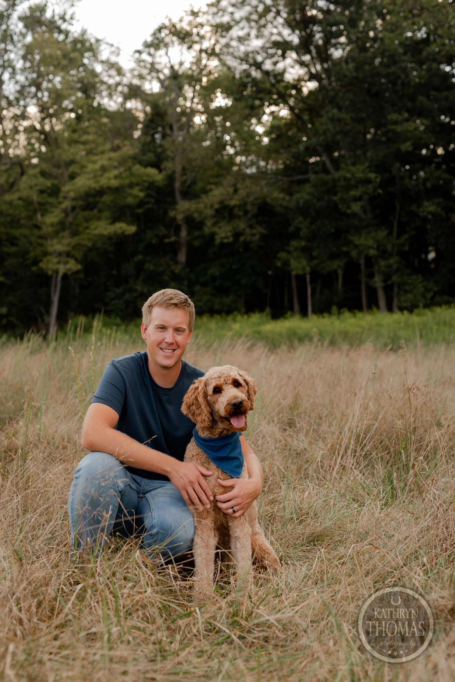 A happy person poses with a goldendoodle dog wearing a blue bandana in a summer meadow.
