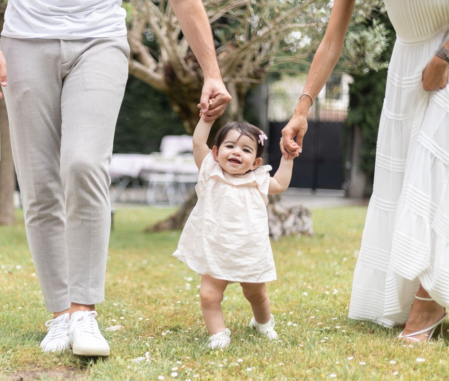 A joyful baby in a white dress learning to walk while holding hands with adults in a sunny garden.