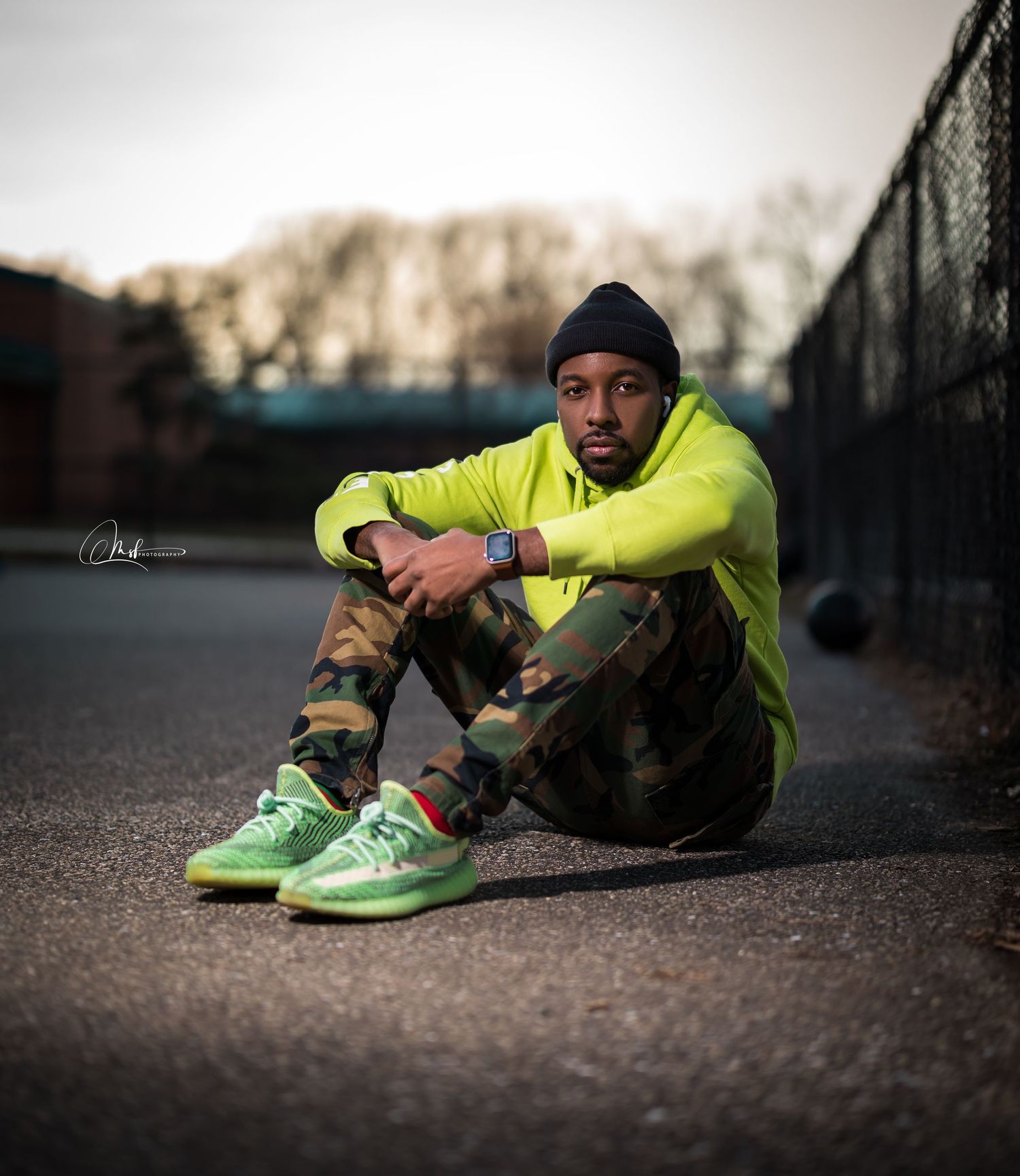 Athletic model in neon yellow hoodie and camo pants sitting on ground with bright green sneakers.