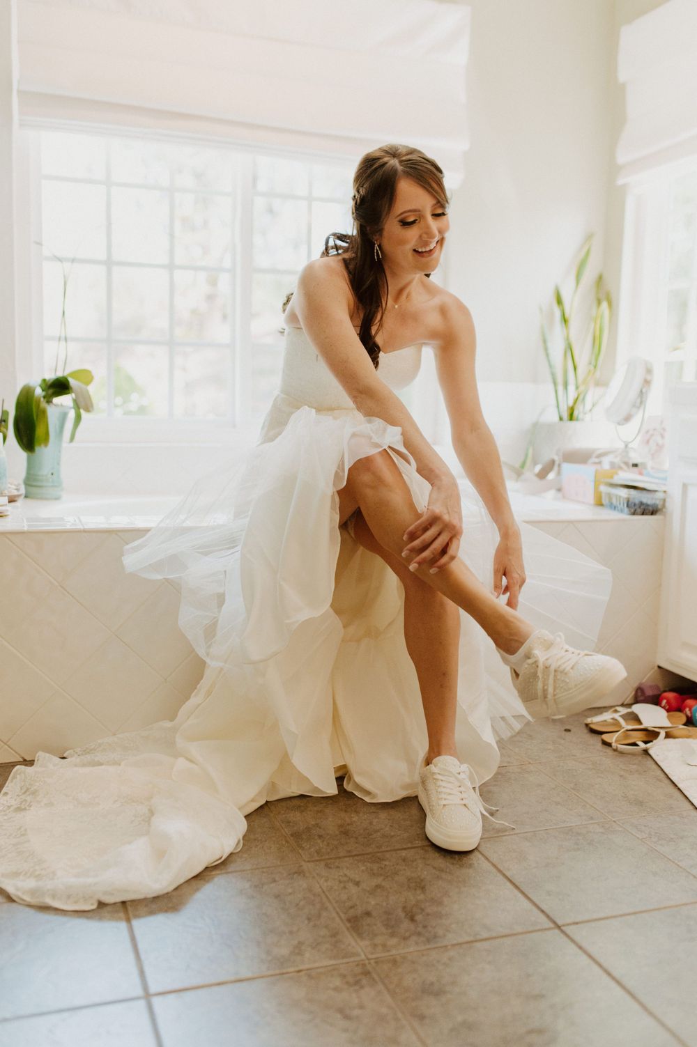 A person in a white wedding dress puts on their shoes while sitting on a tiled floor.
