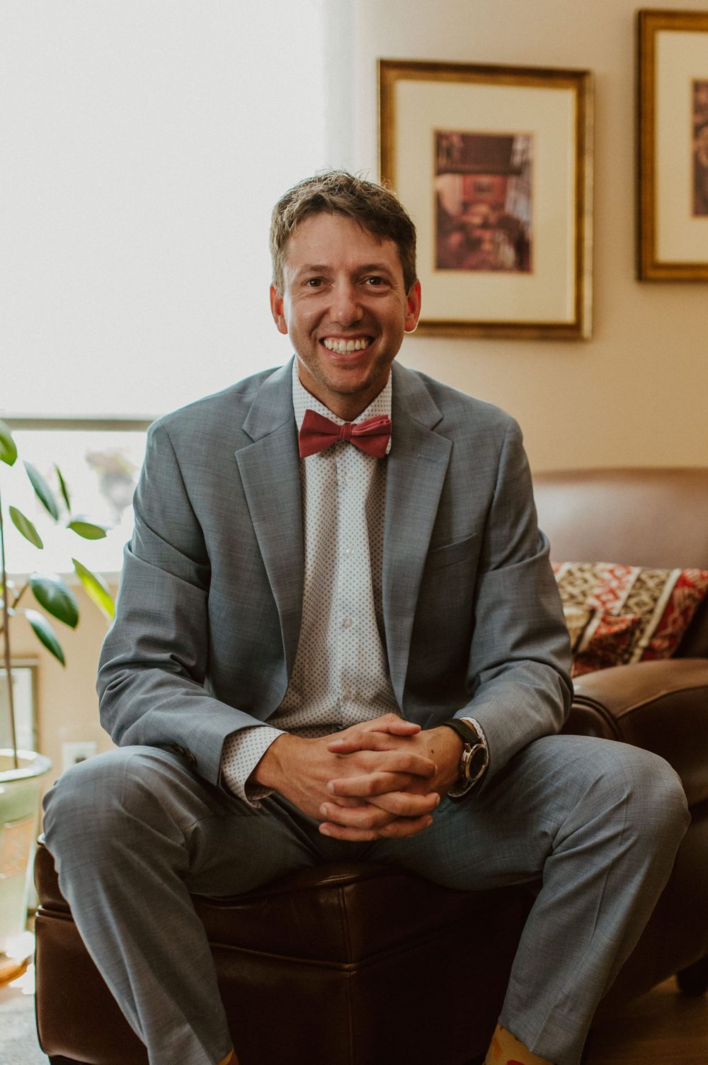 A person in a grey suit and red bowtie sits casually on a leather couch.