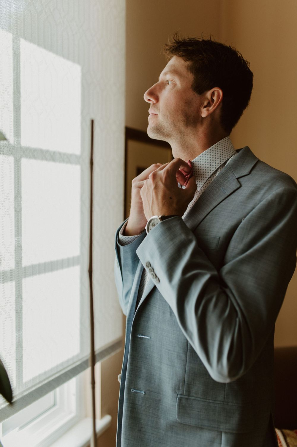 Person in a gray suit jacket looking thoughtfully out a window in warm natural light.