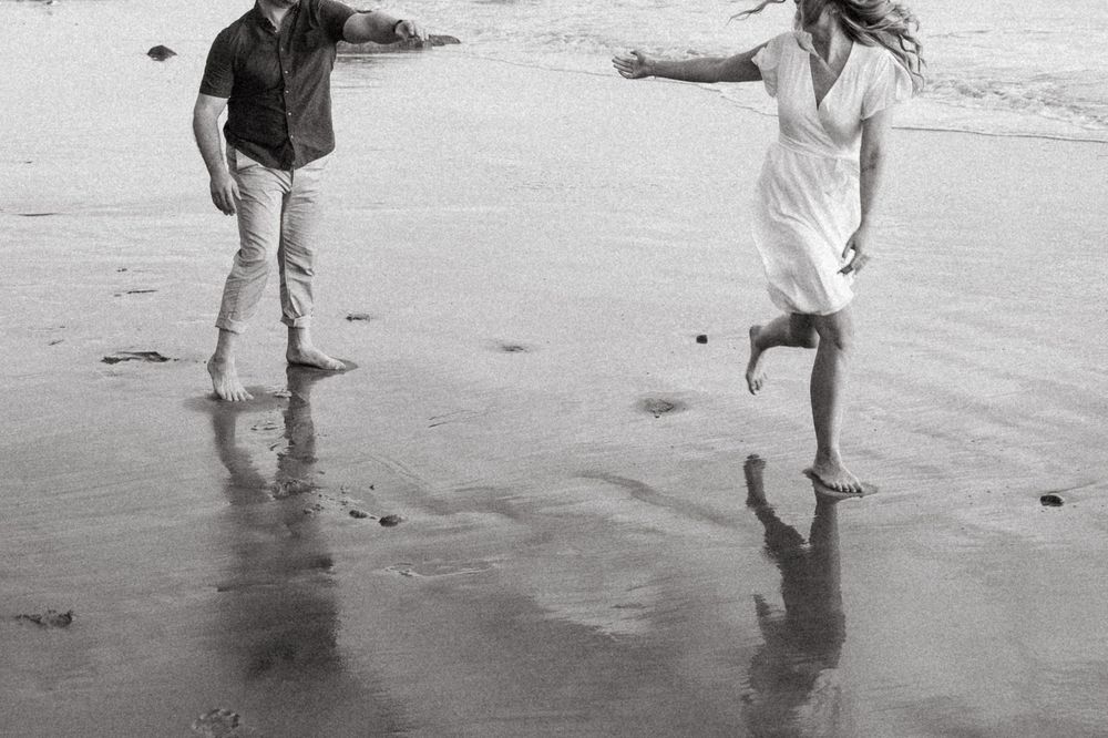 A couple dances playfully together on a wet beach shore in black and white.