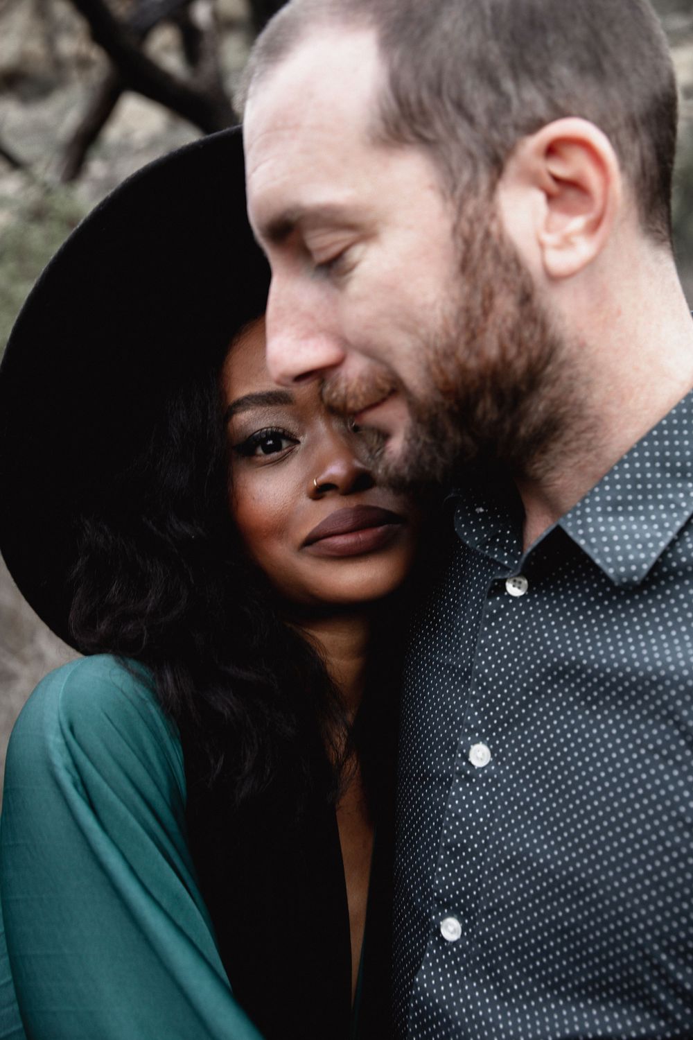A couple in an intimate embrace outdoors, with her wearing a black hat and green top, and him in a polka dot shirt.