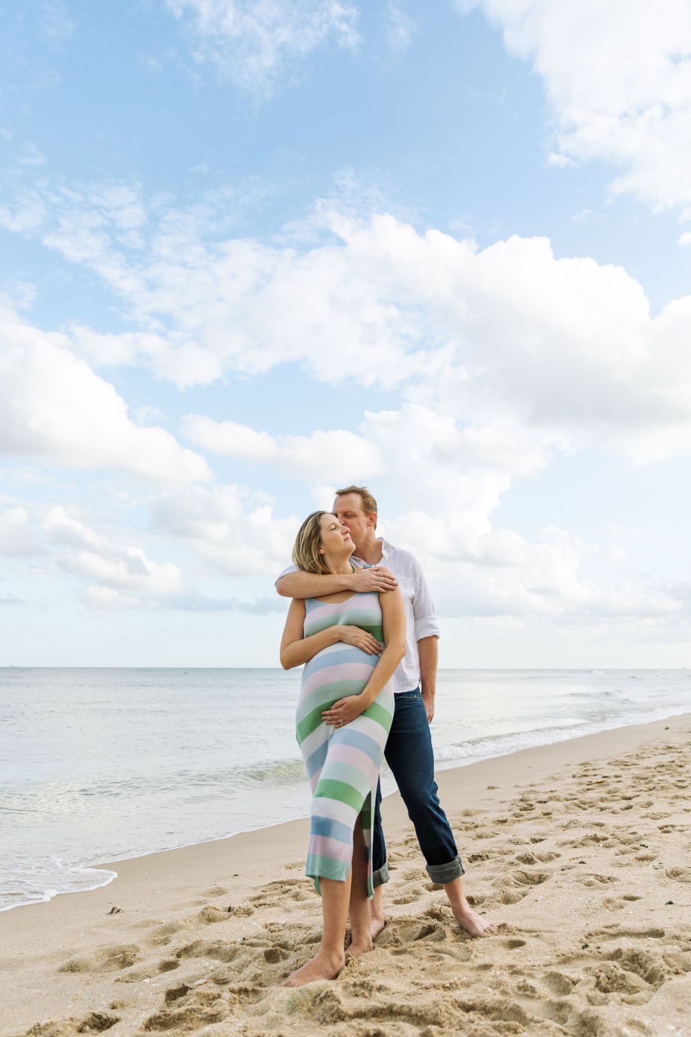 Romantic couple embrace on sandy beach with blue sky and ocean in background, creating serene coastal moment.