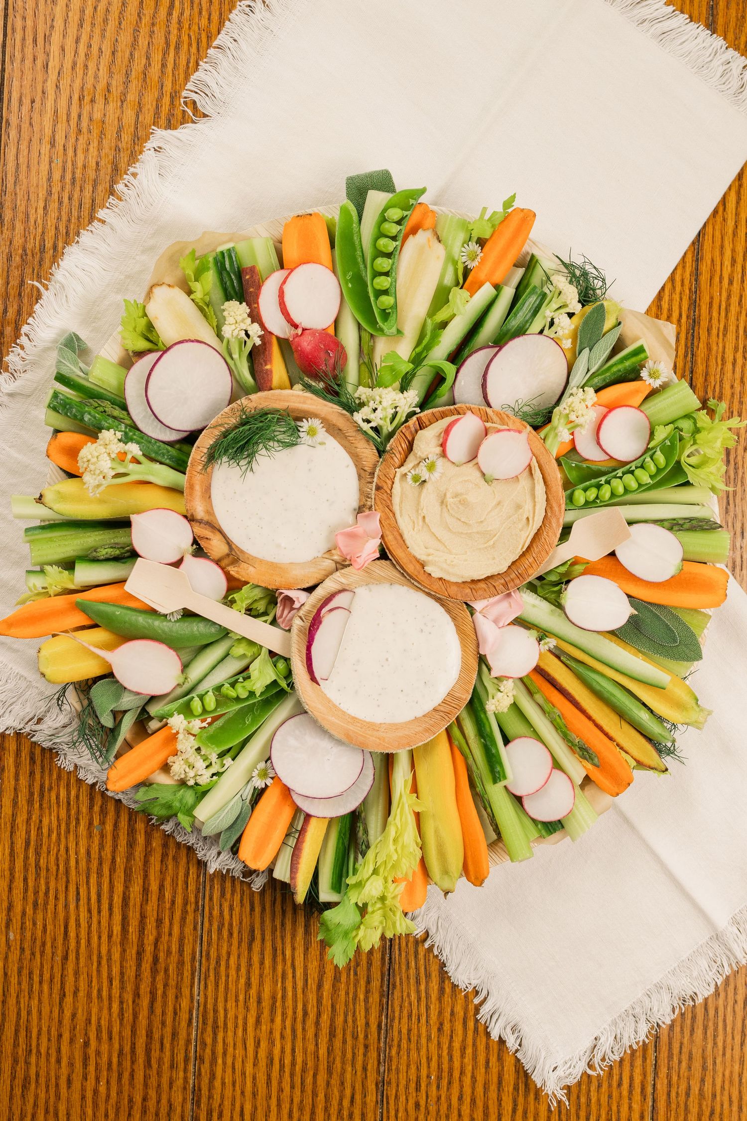 Colorful vegetable crudité platter with assorted dips arranged in a circular pattern on white fabric.