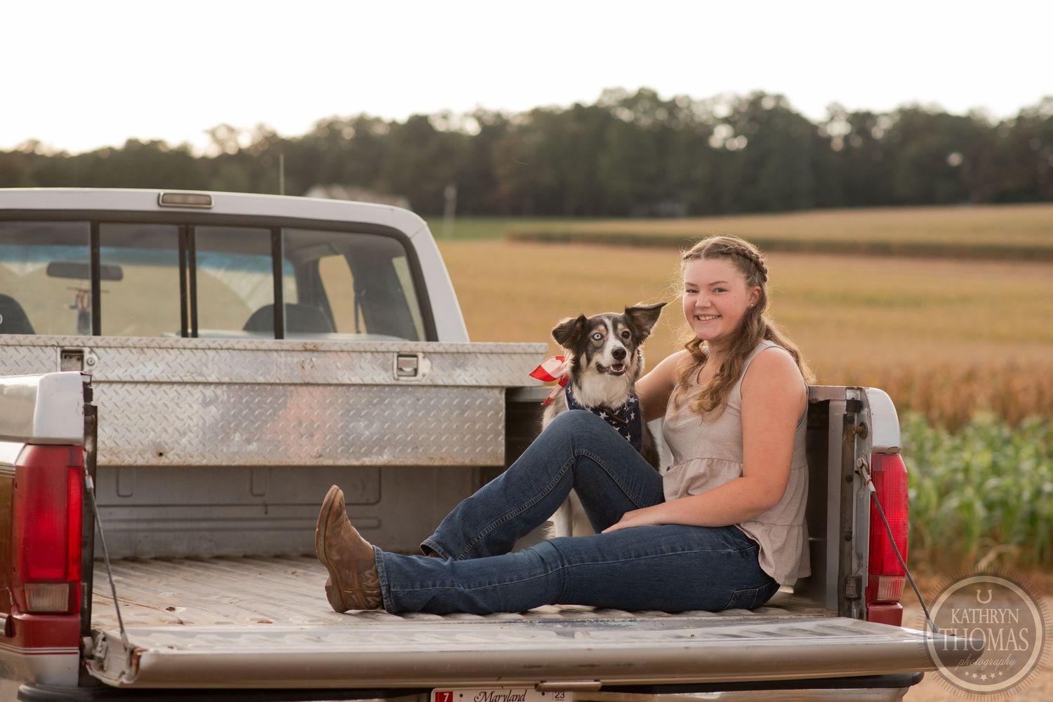Person sitting with dog in pickup truck bed during golden hour with cornfield in background.