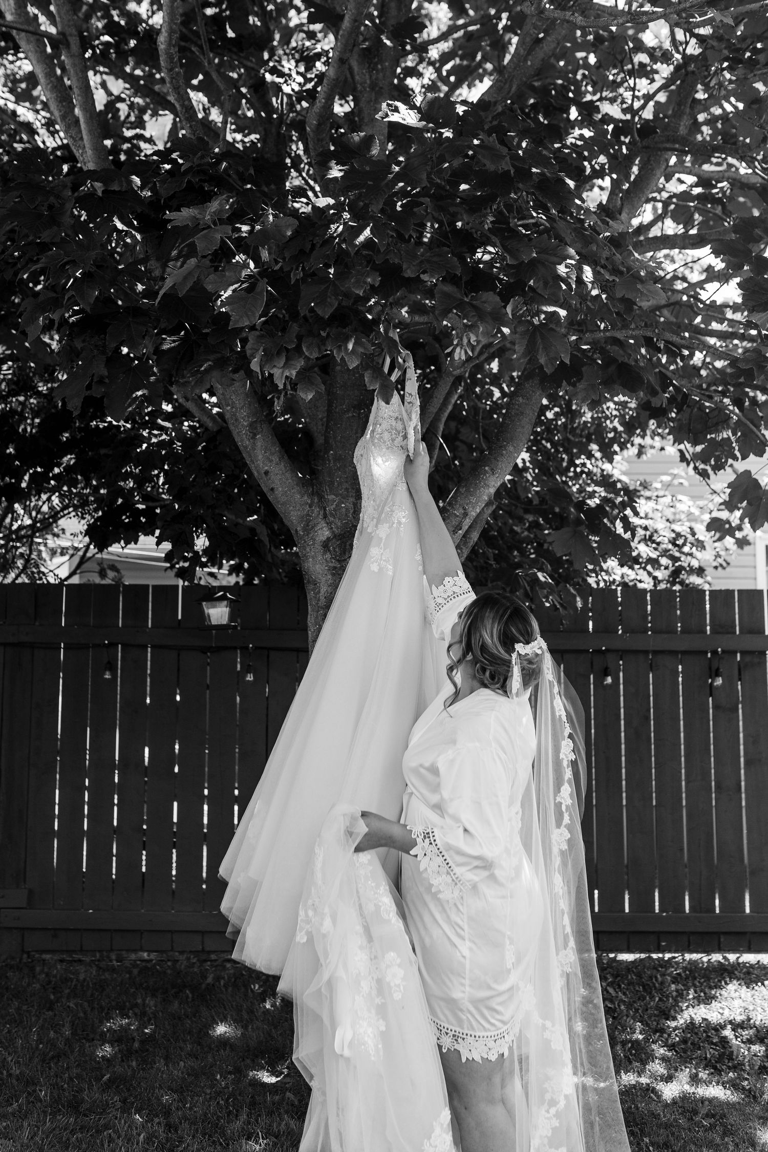 Black and white photo of wedding dress hanging from tree branch against wooden fence.