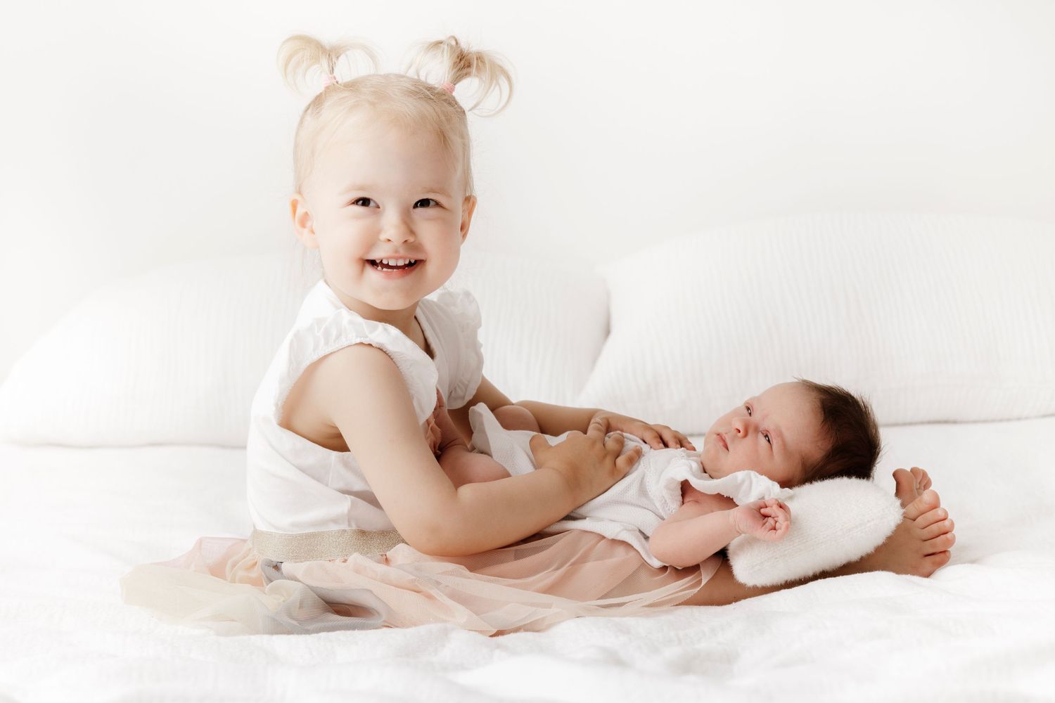 A joyful toddler interacting with a newborn sibling on white bedding.