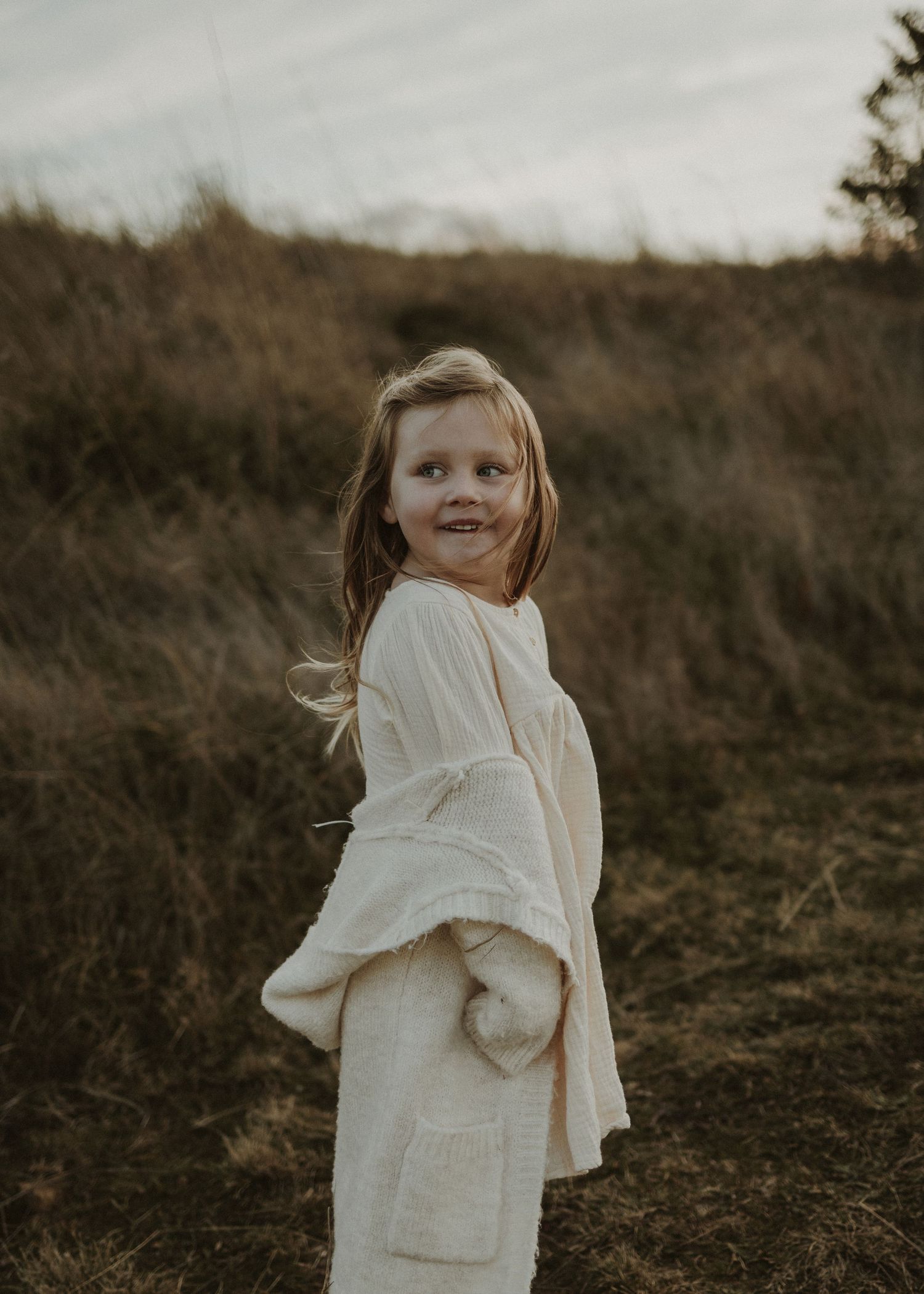 A young girl in white clothing twirls playfully in a field at sunset.