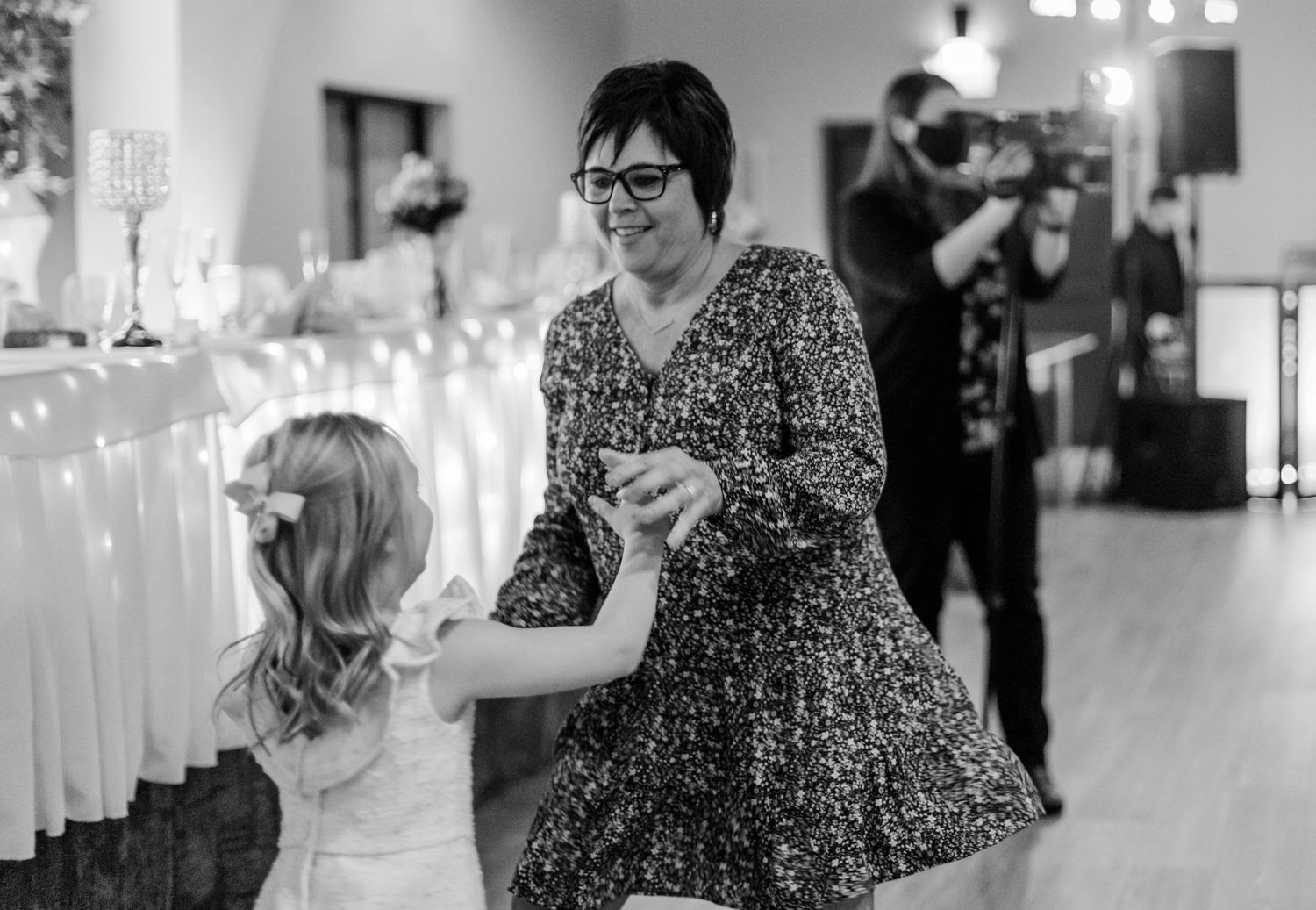 A woman in a patterned dress dances with a young girl at a wedding reception with white decor.