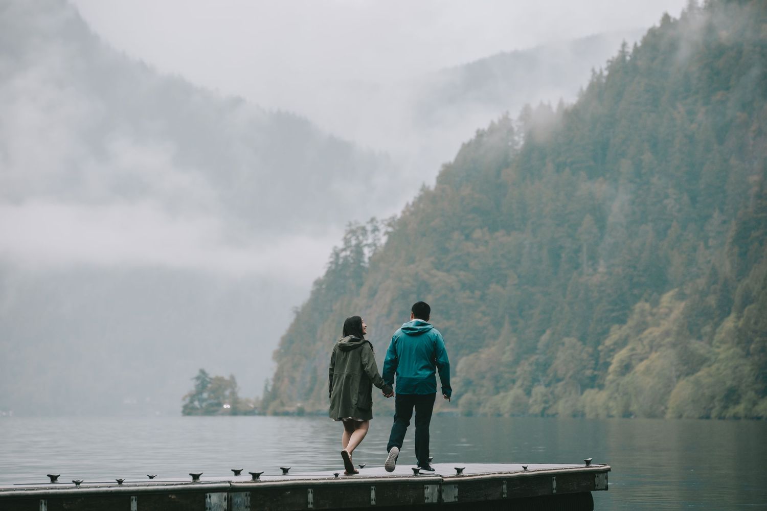 A couple is holding hands walking along a dock at Lake Crescent mountains in the background