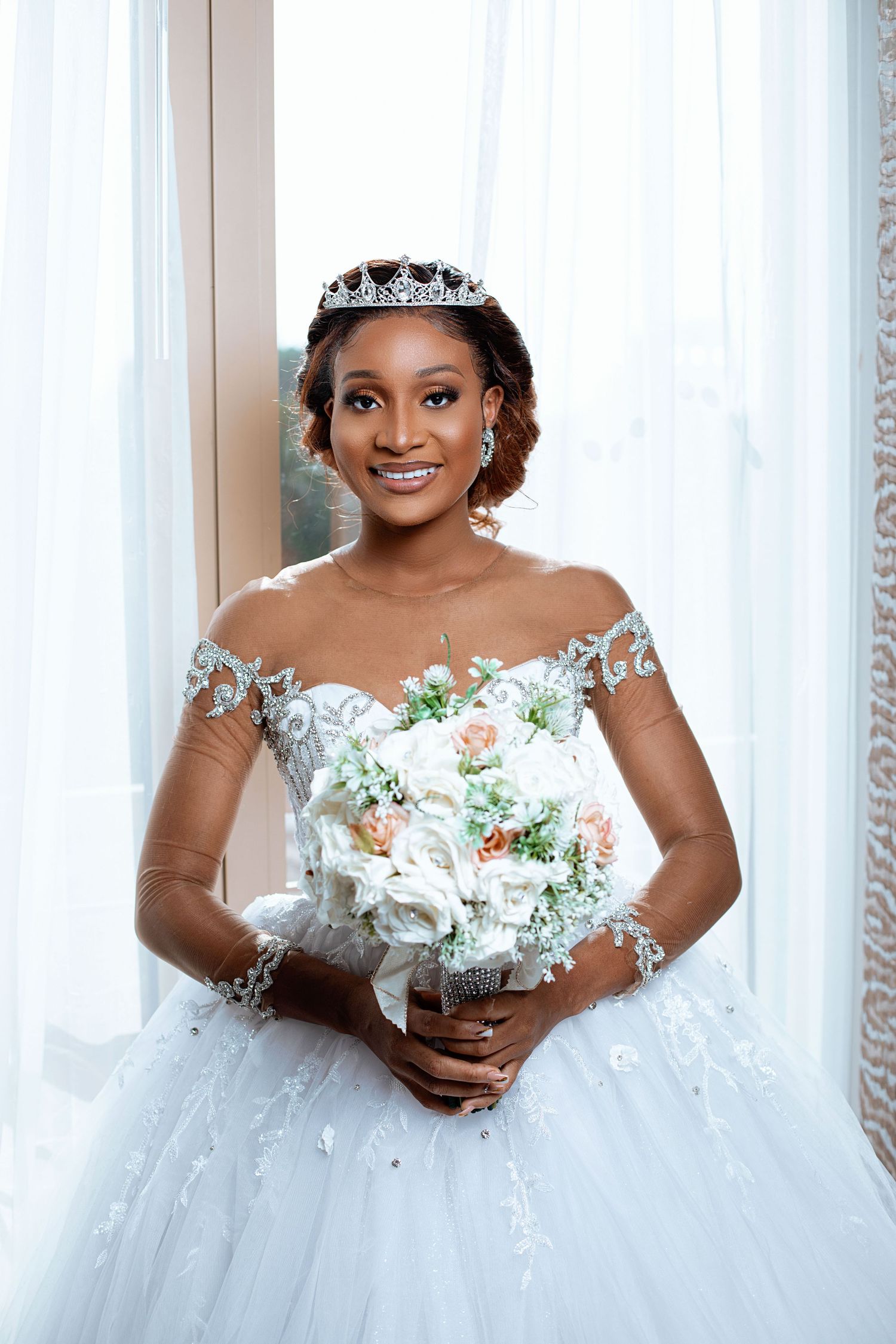 Bride wearing tiara and off-shoulder wedding gown poses with white floral bouquet.