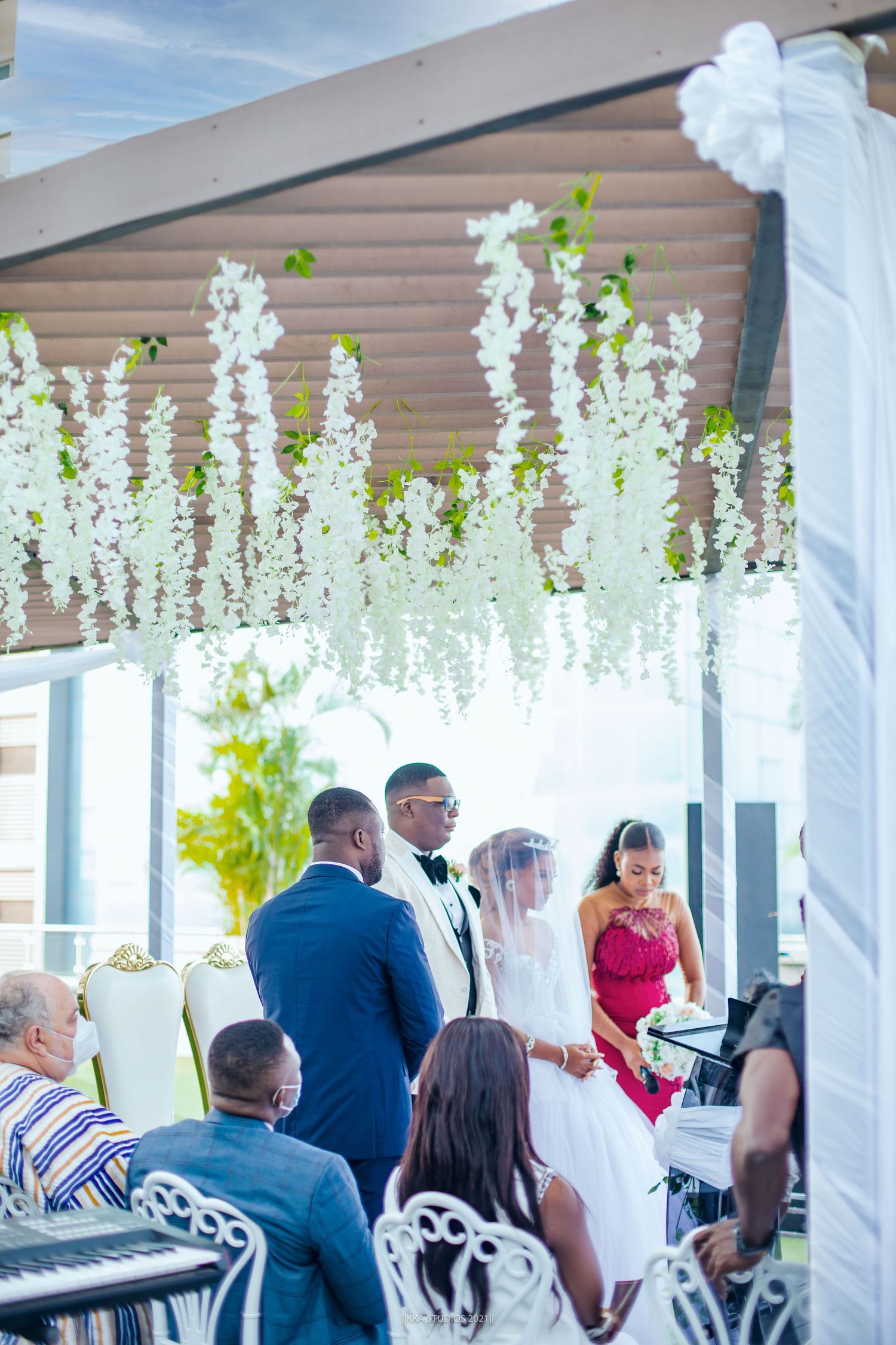 Wedding guests gather under elegant white floral decorations at an outdoor ceremony venue.