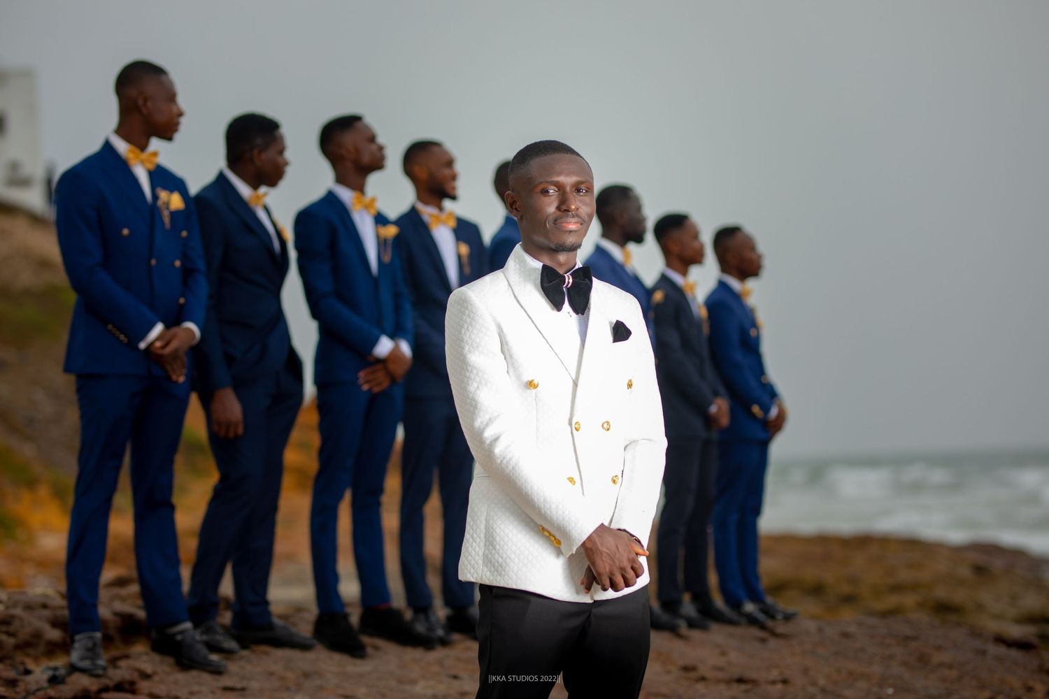 Groomsmen in navy suits and white tuxedo line up for a photo by the ocean.