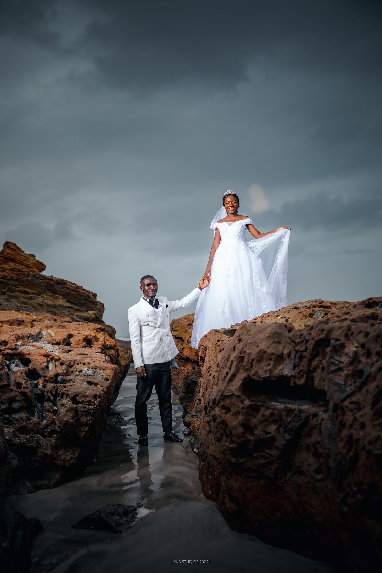 Newlyweds pose for dramatic photos on rocky coastal formation under stormy skies.