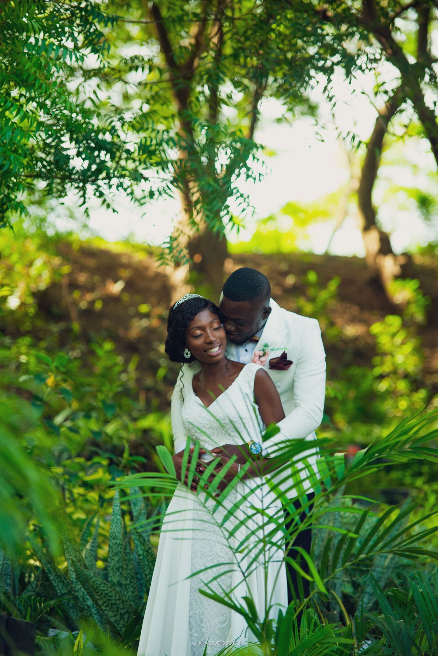 A romantic couple in white outfits poses together against lush tropical greenery and palm fronds.