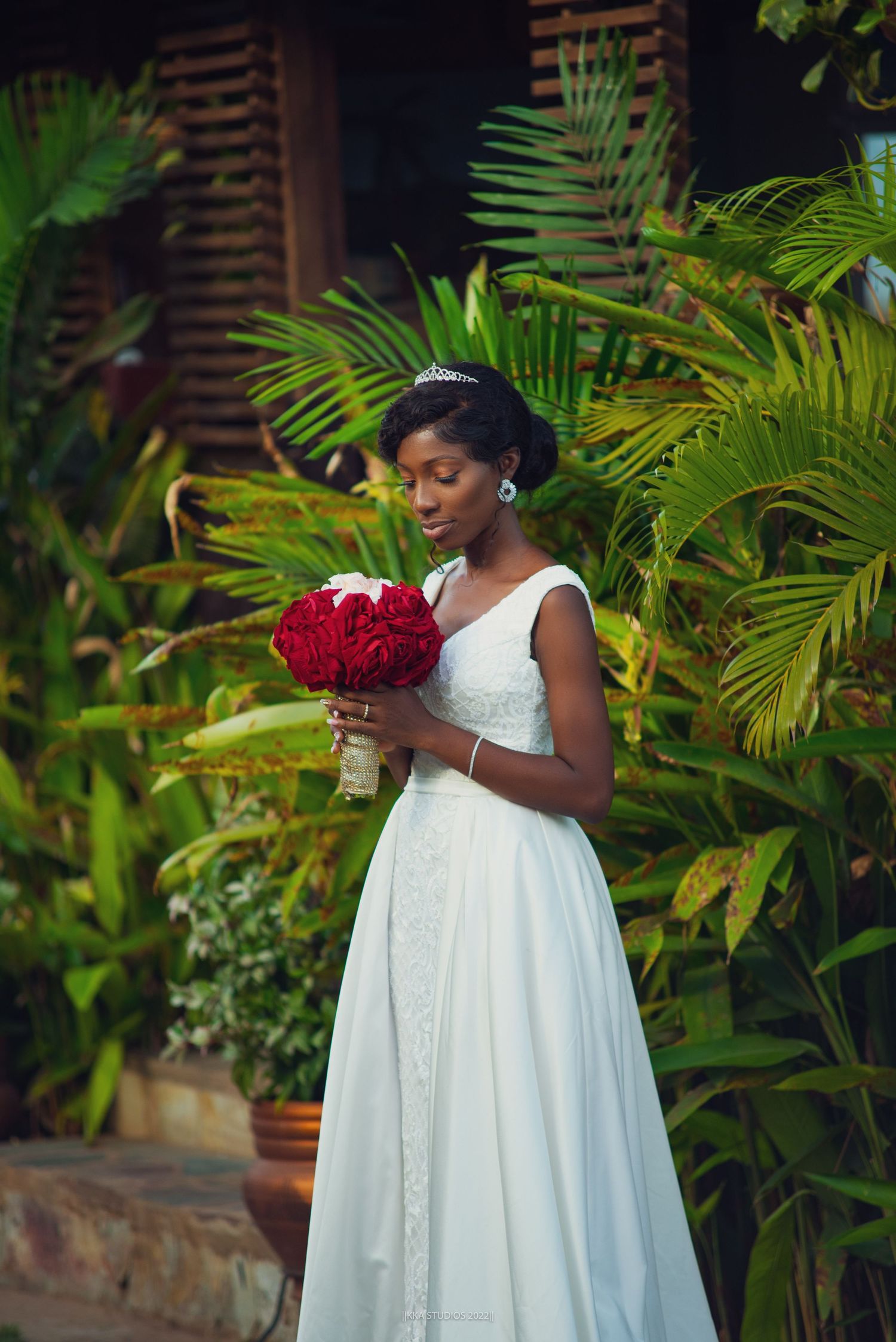 A figure in an elegant white dress stands among tropical plants holding a bouquet of red roses.