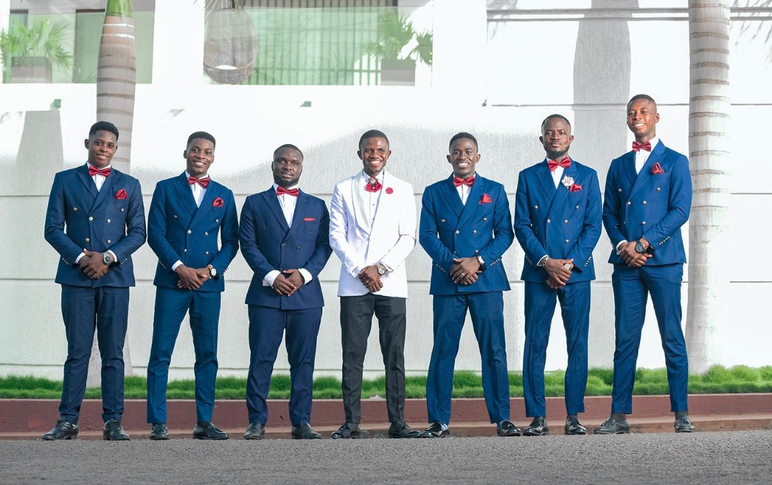 Groomsmen in matching navy suits pose in line against white wall.