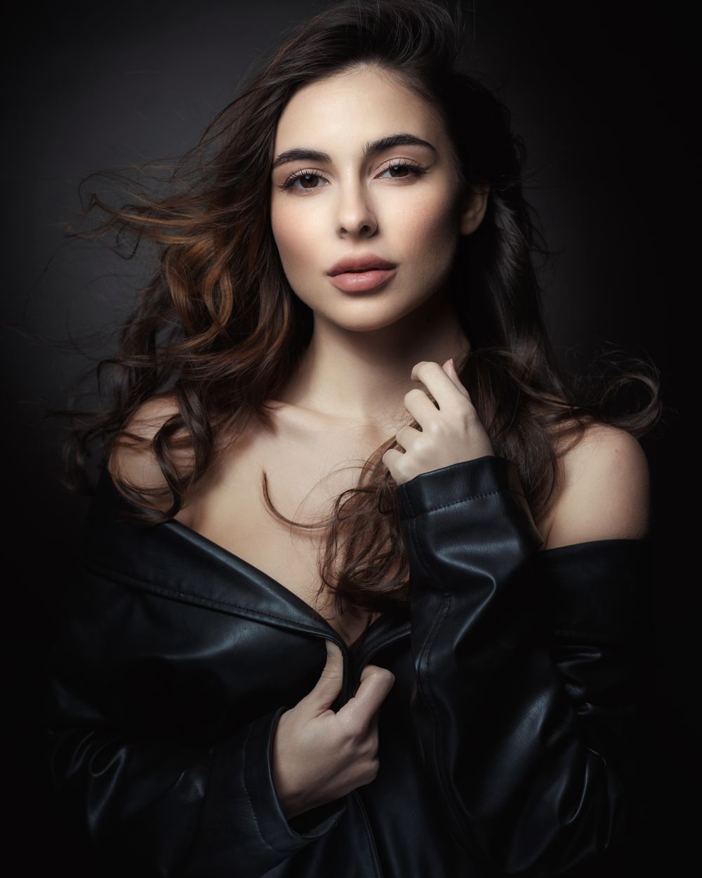 Glamorous beauty portrait with flowing hair and black leather jacket against dark backdrop.