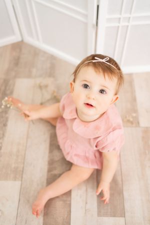 Baby in pink dress sitting on wooden floor looking up with wonder.