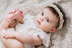 Baby wearing delicate white lace headband lies on soft beige blanket while holding tiny feet.