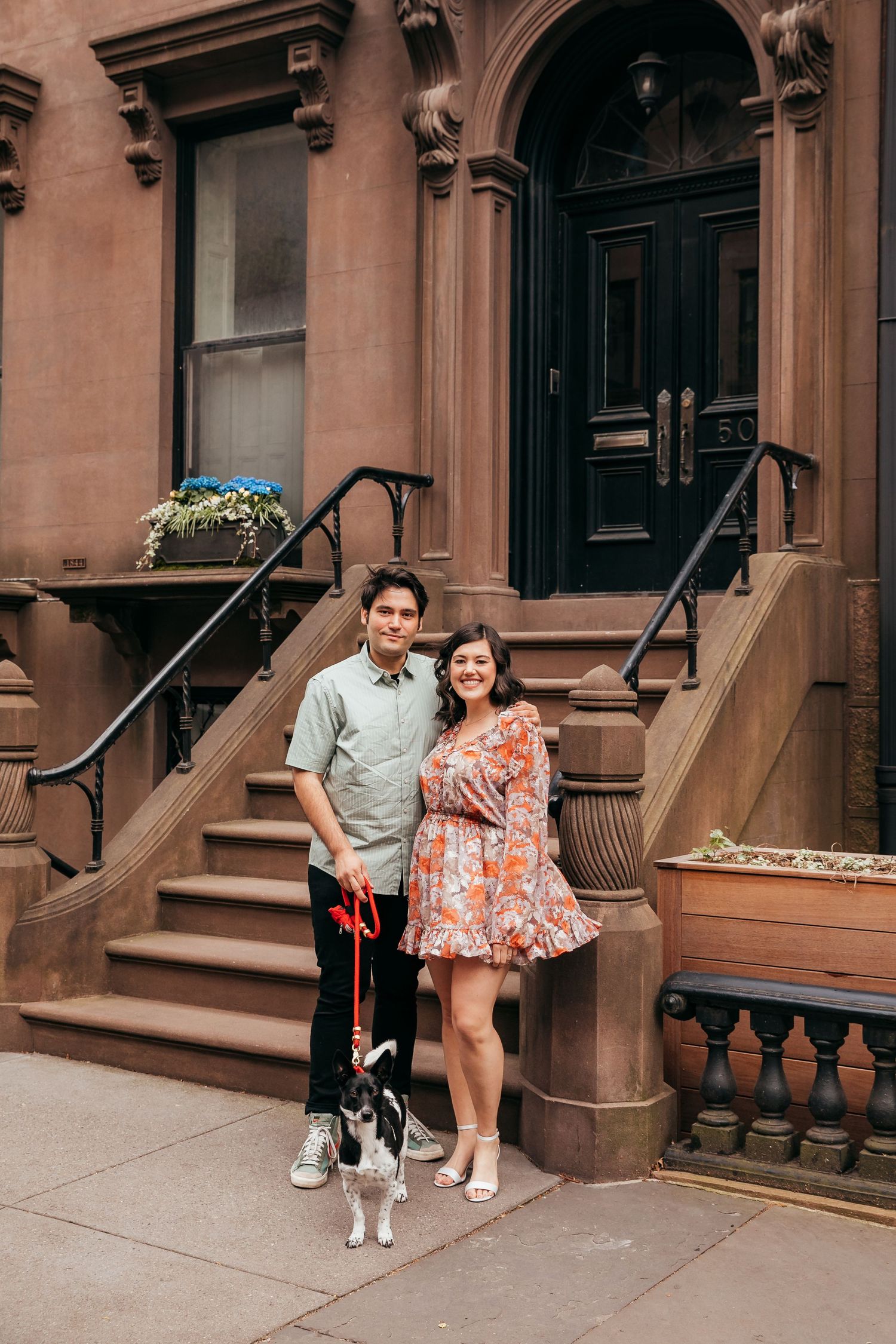 Smiling couple with small dog standing in front of classic brownstone building entrance in New York.